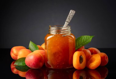 Cam kavanozda kayısı reçeli ve taze meyve. Siyah yansıtıcı arka planda tatlı bir kesişme.