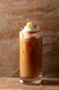 Krem şantili ve karamel soslu buzlu karamel latte, bej seramik masada ferahlatıcı ve tatlı bir kahve..