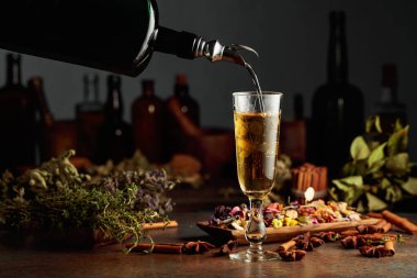 Sarı bitkisel likör ya da karışım eski bir şişeden bardağa dökülür. Kurutulmuş otlar, çiçekler, baharatlar ve eski mutfak aletleri. Bitkisel ilaç kavramı.