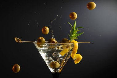 Siyah arka planda yeşil zeytinli, limon kabuklu ve biberiyeli klasik sek martini kokteyli. Sıçratmalı martini. Metnin için boş yer.