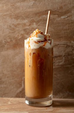 Krem şantili ve karamel soslu buzlu karamel latte, bej seramik masada ferahlatıcı ve tatlı bir kahve..