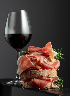 Prosciutto, biberiye ve siyah arka planda bir bardak kırmızı şarap ile ciabatta. Geleneksel İtalyan atıştırması.