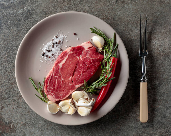 Raw beef steak for grilling with rosemary, garlic, salt, and pepper. Old stone background. Top view.