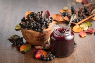 Chokeberry jam and fresh berries with leaves on an old wooden table. clipart