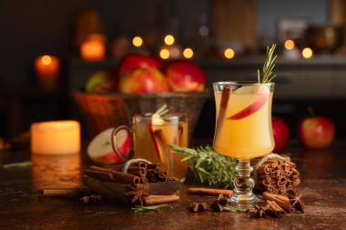 Elma dilimleri, tarçın, biberiye ve kahverengi bir kırsal masada cam bardakta anason şarabı. Karanlık bir arka planda mutfak aletleri ve yanan mumlar..