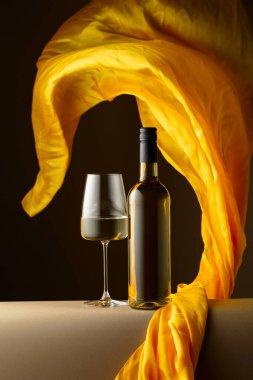 Şişe ve bir bardak beyaz şarap ve koyu arkaplanda çırpınan sarı kumaş..