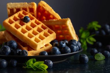 Siyah tabakta taze yaban mersinli Belçika waffleları. Koyu mavi bir masada yeşil yapraklı waffle ve taze meyveler..