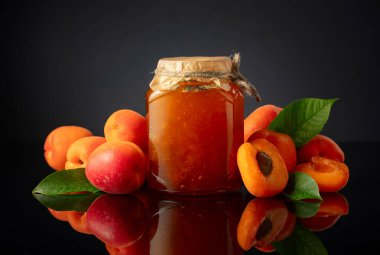 Cam kavanozda kayısı reçeli ve taze meyve. Siyah yansıtıcı arka planda tatlı bir kesişme.