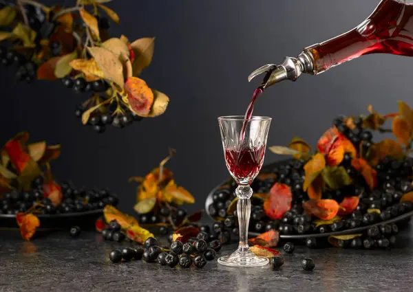 stock image Black chokeberry liqueur and fresh berries on a black stone table. Sweet flavour drink is poured from a bottle into a glass.