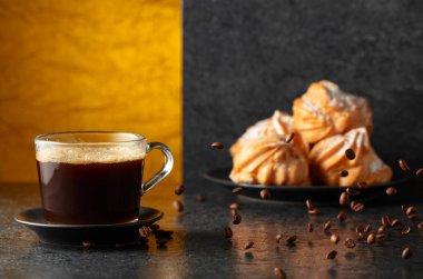 Cam bardakta sade kahve ve siyah masada hareketli kahve çekirdekleri. Arka plandaki kremalı keklerin içinde. Boşluğu kopyala.