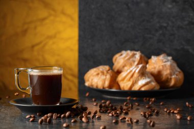 Cam bardakta sade kahve ve siyah masada hareketli kahve çekirdekleri. Arka plandaki kremalı keklerin içinde. Boşluğu kopyala.