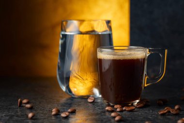 Siyah taş bir masada espresso ve bir bardak soğuk su. Kavrulmuş kahve çekirdekleri hareket halinde. Boşluğu kopyala.