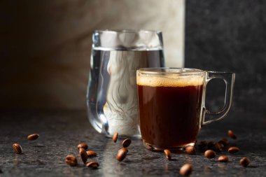 Siyah taş bir masada espresso ve bir bardak soğuk su. Kavrulmuş kahve çekirdekleri hareket halinde. Boşluğu kopyala.