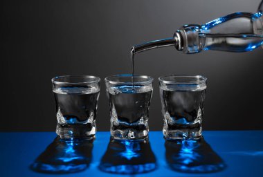 Votka şişeden koyu bir arka planda bir bardağa dökülüyor. Mavi arka ışık. Boşluğu kopyala.