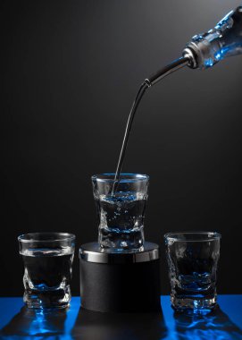 Votka şişeden koyu bir arka planda bir bardağa dökülüyor. Mavi arka ışık. Boşluğu kopyala.