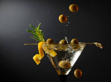 Siyah arka planda yeşil zeytinli, limon kabuklu ve biberiyeli klasik sek martini kokteyli. Sıçratmalı martini. Metnin için boş yer.