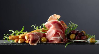 Siyah arka planda ekmek çubuğu, zeytin ve biberiyeli jambon. Geleneksel İtalyan atıştırmaları.