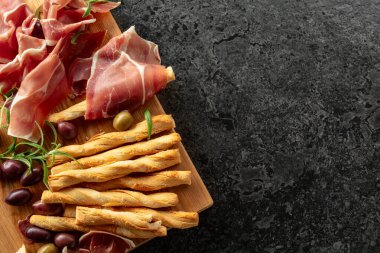 Tahta kesme tahtasında ekmek çubuğu, zeytin ve biberiyeli jambon. Geleneksel İtalyan atıştırmalıkları siyah taş bir masada..