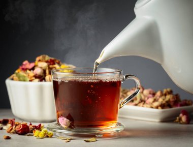 Sıcak bitkisel çay bardağa dökülür. Kurutulmuş otlar ve çiçekler masanın üzerine saçılmış..