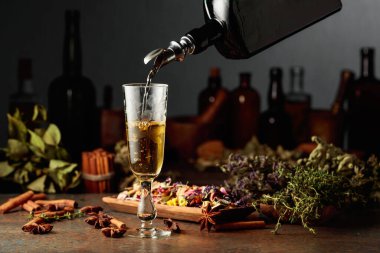 Sarı bitkisel likör ya da karışım eski bir şişeden bardağa dökülür. Kurutulmuş otlar, çiçekler, baharatlar ve eski mutfak aletleri. Bitkisel ilaç kavramı.