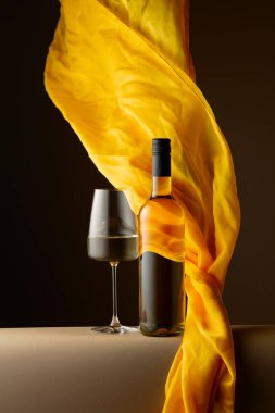 Şişe ve bir bardak beyaz şarap ve koyu arkaplanda çırpınan sarı kumaş..