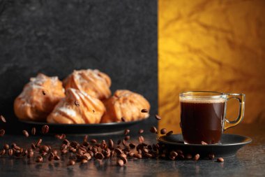 Cam bardakta sade kahve ve siyah masada hareketli kahve çekirdekleri. Arka plandaki kremalı keklerin içinde. Boşluğu kopyala.