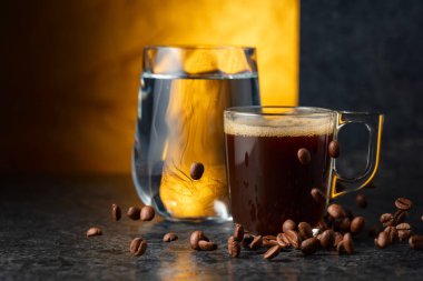 Siyah taş bir masada espresso ve bir bardak soğuk su. Kavrulmuş kahve çekirdekleri hareket halinde. Boşluğu kopyala.