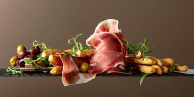 Ekmek çubuğu, zeytin ve biberiyeli jambon. Kahverengi arka planda geleneksel İtalyan atıştırmalıkları.