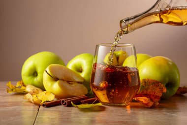 Elma suyu şişeden bardağa dökülür. Seramik bir masada olgun elmalar, tarçın çubukları ve kurumuş yapraklar. Kopyalama alanı olan bej arkaplan.