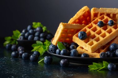Siyah tabakta taze yaban mersinli Belçika waffleları. Koyu mavi bir masada yeşil yapraklı waffle ve taze meyveler..