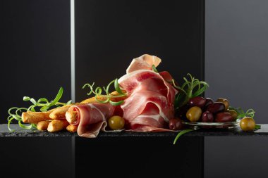 Siyah arka planda ekmek çubuğu, zeytin ve biberiyeli jambon. Geleneksel İtalyan atıştırmaları.