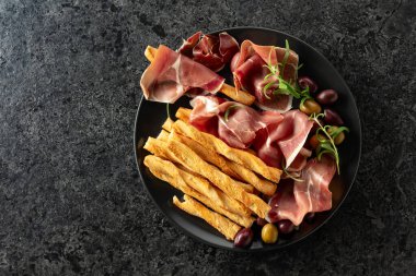 Siyah tabakta ekmek çubuğu, zeytin ve biberiyeli jambon. Geleneksel İtalyan atıştırmalıkları siyah taş bir masada. Üst görünüm.