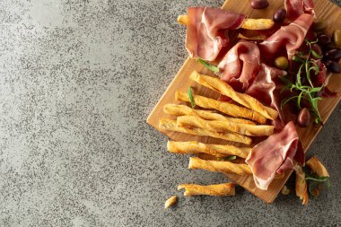 Tahta kesme tahtasında ekmek çubuğu, zeytin ve biberiyeli jambon. Gri taş bir masada geleneksel İtalyan atıştırmalıkları..