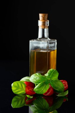 Siyah yansıtıcı arka planda fesleğenli bir şişe zeytinyağı ve domates..