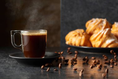 Cam bardakta sade kahve ve siyah masada hareketli kahve çekirdekleri. Arka plandaki kremalı keklerin içinde. Boşluğu kopyala.