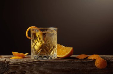 Kısa kristal bardakta zengin bir Old Fashioned kokteyli, buzlu koyu kehribar likörü, eski bir tahta üzerinde portakal kabuğu ile süslenmiş. Boşluğu kopyala.