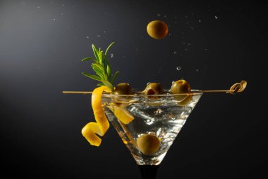 Siyah arka planda yeşil zeytinli, limon kabuklu ve biberiyeli klasik sek martini kokteyli. Sıçratmalı martini. Metnin için boş yer.