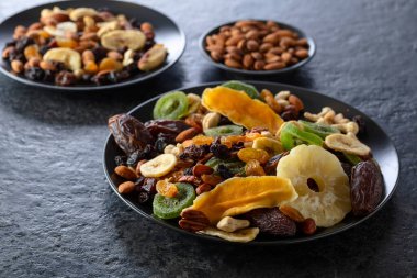 Kurumuş tropikal meyveler, fındıklar ve kuru üzümler siyah bir taş masada..