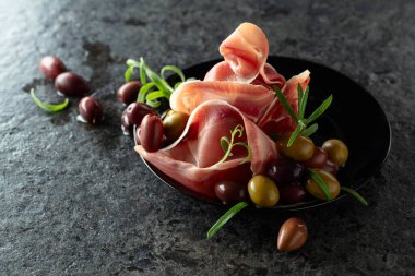 Siyah taş bir masada zeytin ve biberiyeli İtalyan jambonu. Boşluğu kopyala.