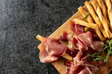 Tahta kesme tahtasında ekmek çubuğu, zeytin ve biberiyeli jambon. Geleneksel İtalyan atıştırmalıkları siyah taş bir masada..
