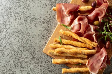 Tahta kesme tahtasında ekmek çubuğu, zeytin ve biberiyeli jambon. Gri taş bir masada geleneksel İtalyan atıştırmalıkları..