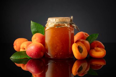 Cam kavanozda kayısı reçeli ve taze meyve. Siyah yansıtıcı arka planda tatlı bir kesişme.