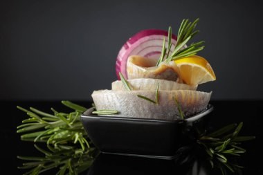 Limonlu, kırmızı soğanlı ve biberiyeli ringa fileto. Siyah yansıtıcı arka planda ringa balığı olan siyah bir tabak. Boşluğu kopyala.