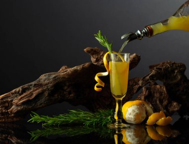 Siyah yansıtıcı arka planda geleneksel İtalyan likörü Limoncello. Bir bardak limon likörü ve arka planda biberiye dalı. İçki bardağa dökülür..