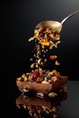 Çeşitli kurutulmuş otlar ve baharatlar ahşap bir kaba dökülür. Siyah yansıtıcı arkaplan.