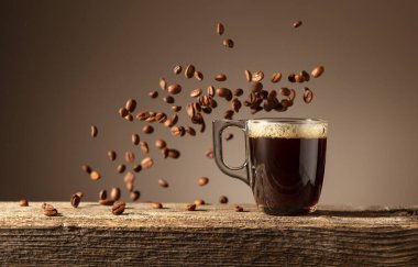 Eski ahşap bir masada cam bardakta espresso. Kavrulmuş kahve çekirdekleri hareket halinde. Boşluğu kopyala.