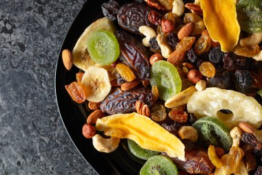 Kurumuş tropikal meyveler, fındıklar ve kuru üzümler siyah bir taş masada..