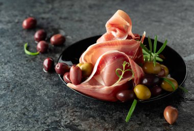 Siyah taş bir masada zeytin ve biberiyeli İtalyan jambonu. Boşluğu kopyala.