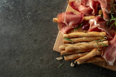 Tahta kesme tahtasında ekmek çubuğu, zeytin ve biberiyeli jambon. Taş bir masada geleneksel İtalyan atıştırmalıkları.