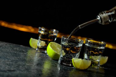 Tekila shot bardağına dökülür. Siyah arka planda alkollü içecek ve limon dilimleri. Boşluğu kopyala.